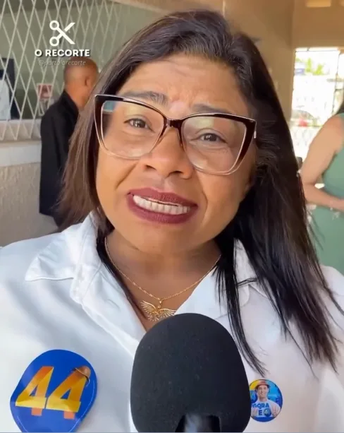 Nilda Cruz, de óculos e camisa branca com adesivos de campanha, fala diante de um microfone