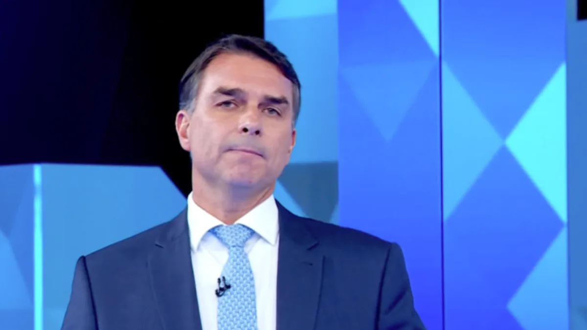 Flávio Bolsonaro discursando em púlpito com logo da Globo ao fundo.