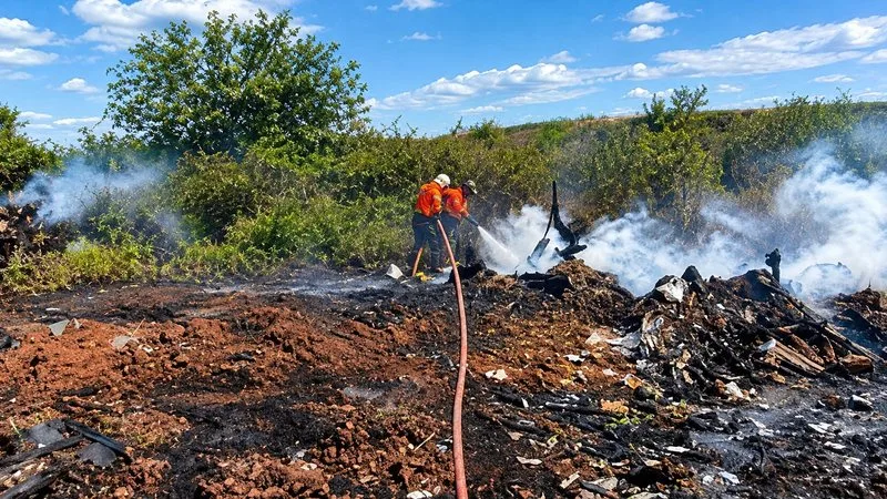 Caminhões de combate a incêndio atuando em área de vegetação com fumaça ao fundo.