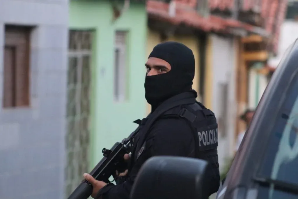 Policial com uniforme tático e arma longa durante operação em área urbana.