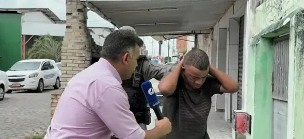 Homem sendo entrevistado por repórter na presença de um policial militar após sua prisão.