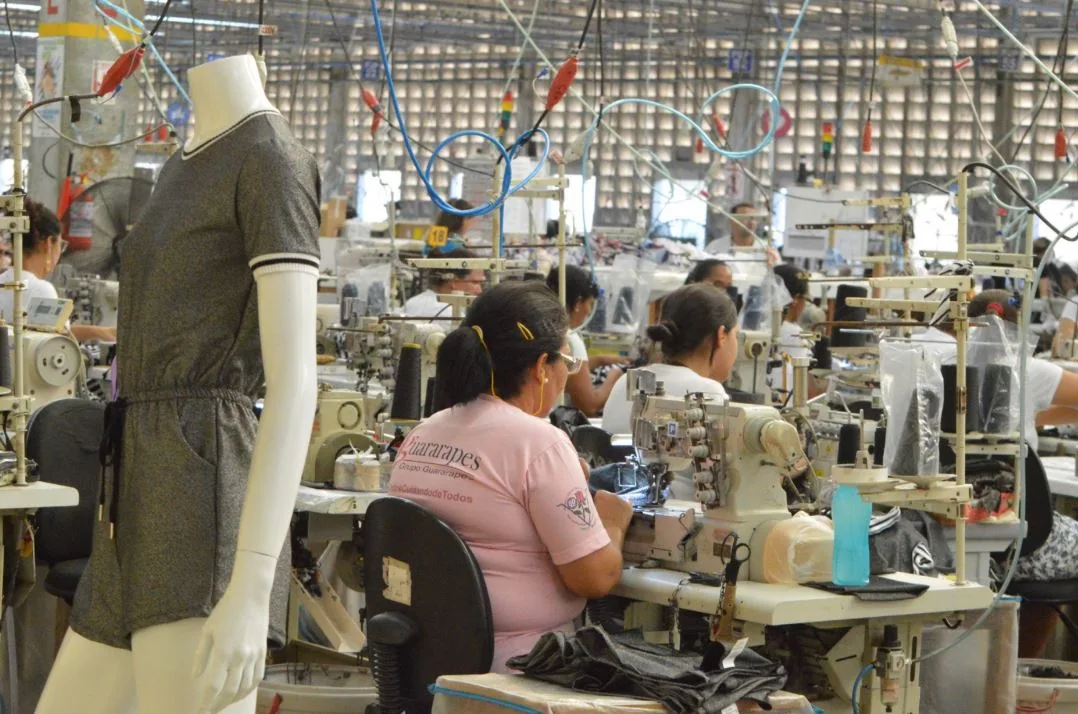 Operárias trabalhando em máquinas de costura dentro de uma fábrica têxtil.