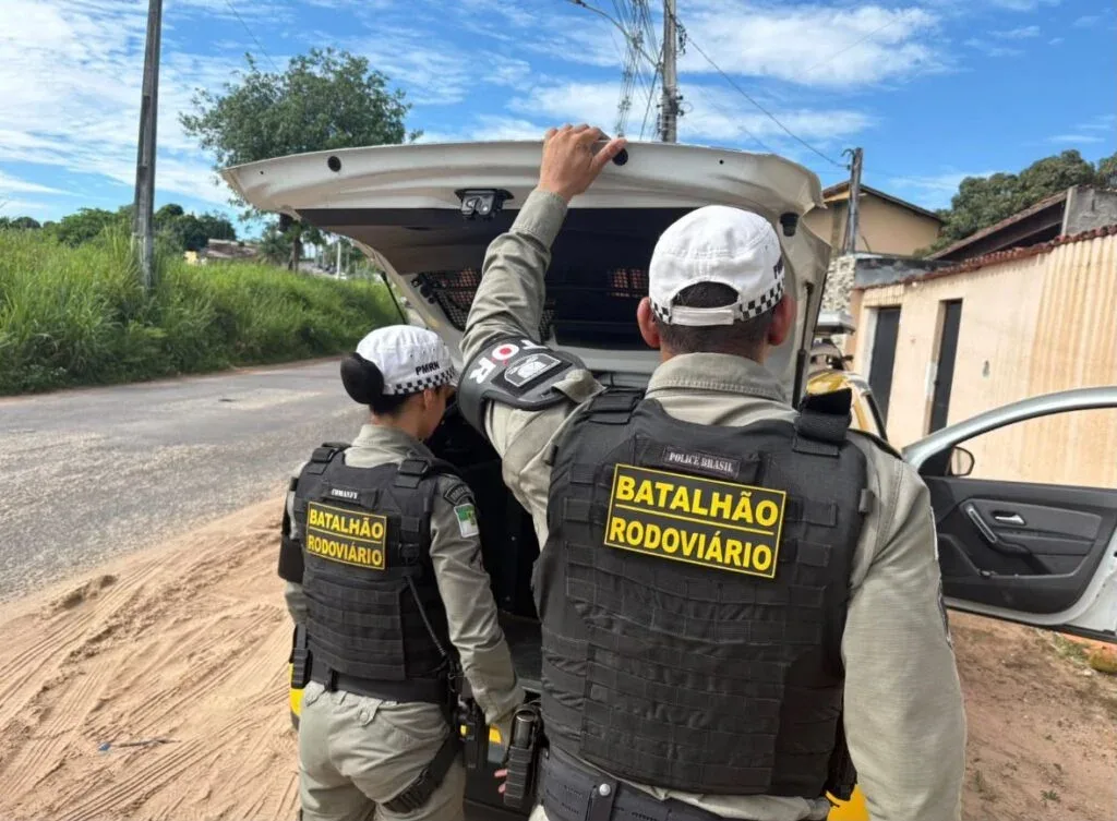 Dois policiais militares do Batalhão Rodoviário, vistos de costas e usando coletes de proteção, estão junto ao compartimento traseiro aberto de uma viatura estacionada em uma rua.