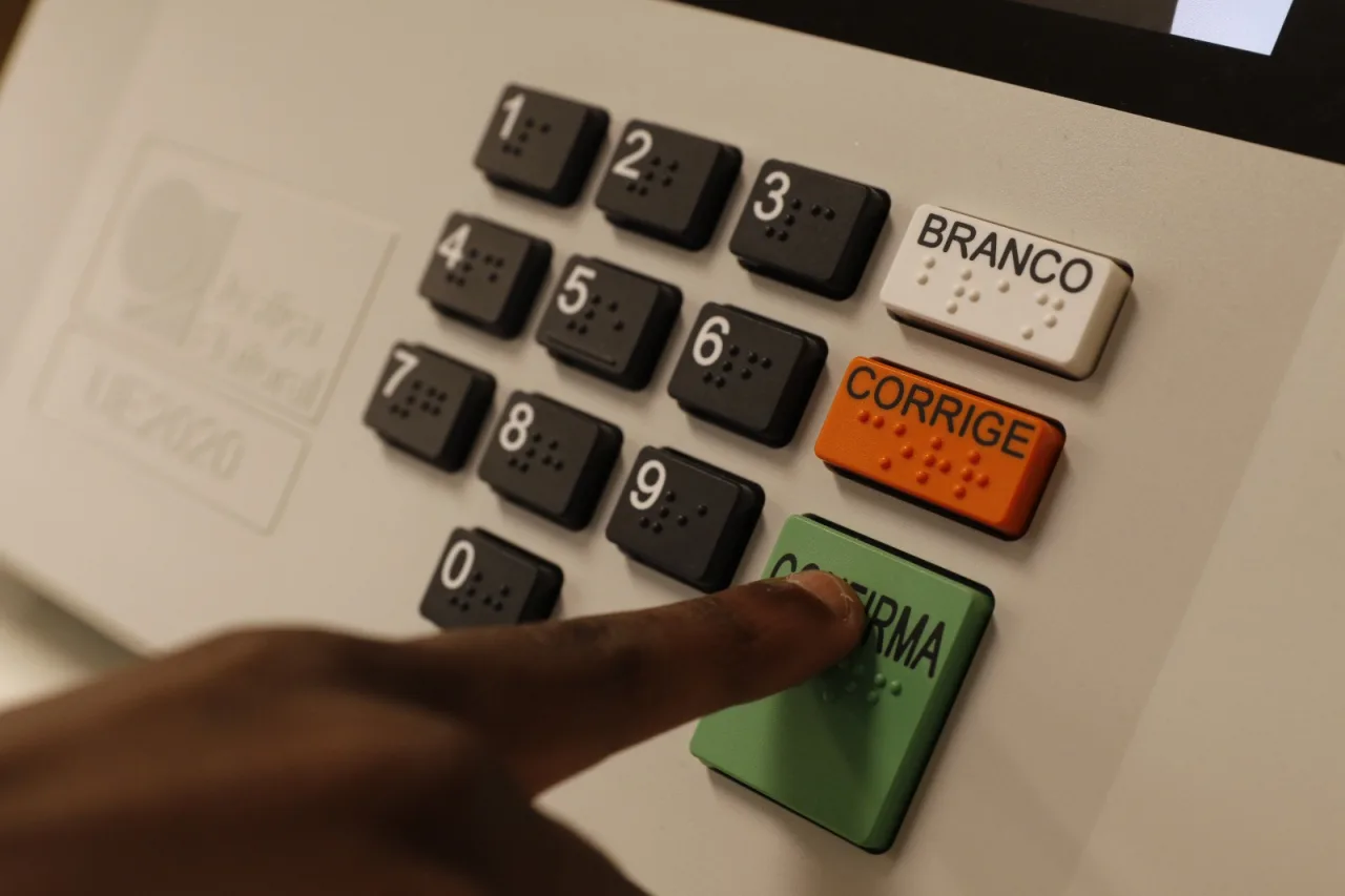 Close de urna eletrônica com teclado numérico e teclas branco, corrige e confirma, enquanto eleitor pressiona o botão verde