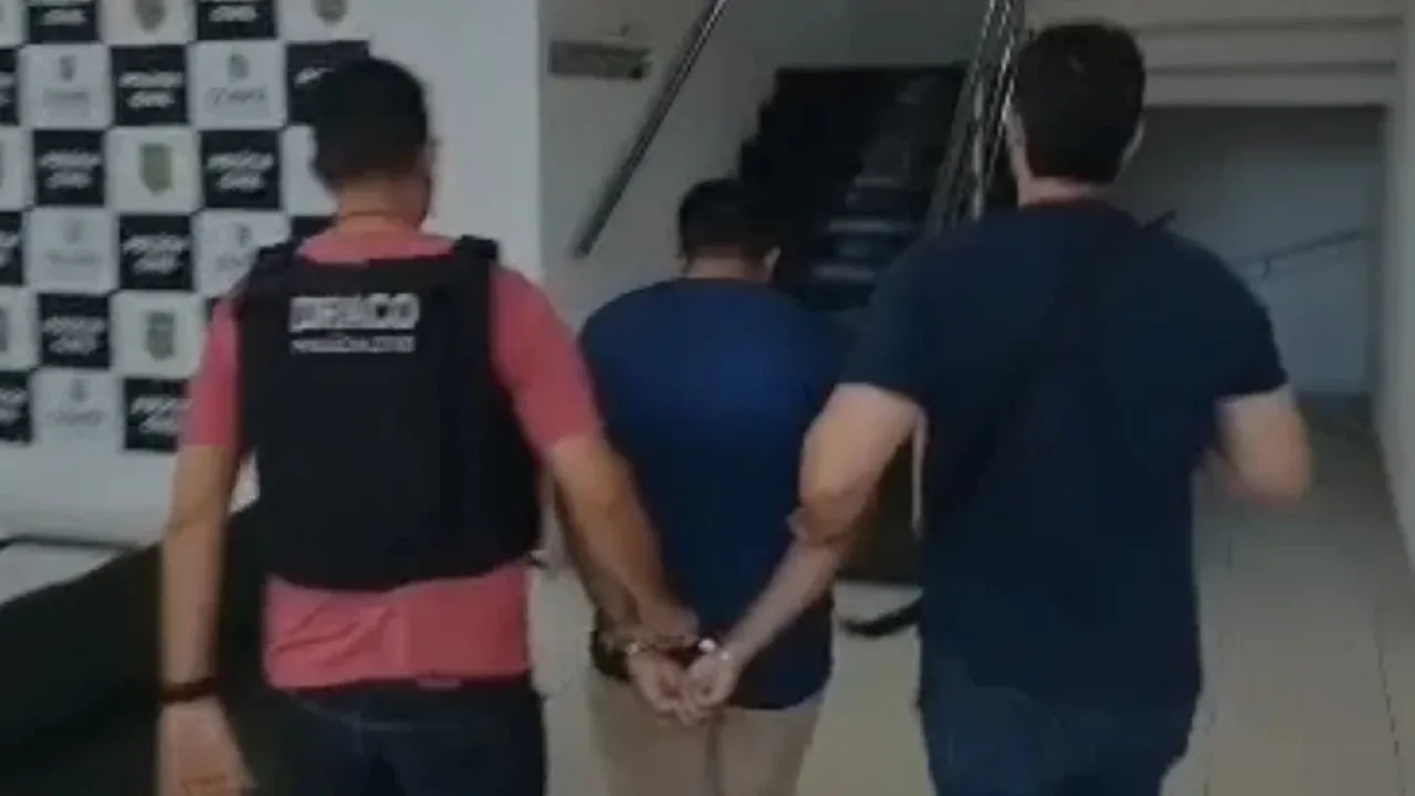 Homem de camisa azul sendo conduzido por policiais em operação contra facção criminosa.