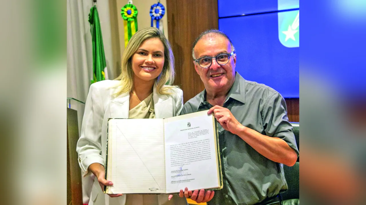 Joanna Guerra e Paulinho Freire sorridentes em frente a bandeiras institucionais.