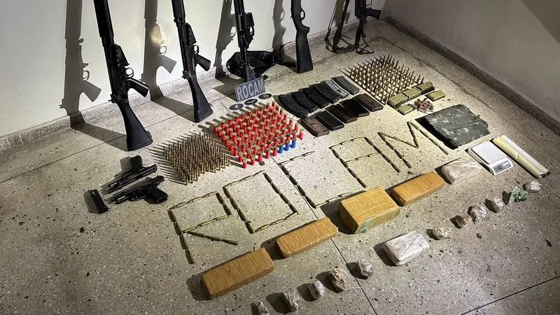 Mesa com fuzis, pistolas e porções de drogas apreendidas pela Polícia Militar.