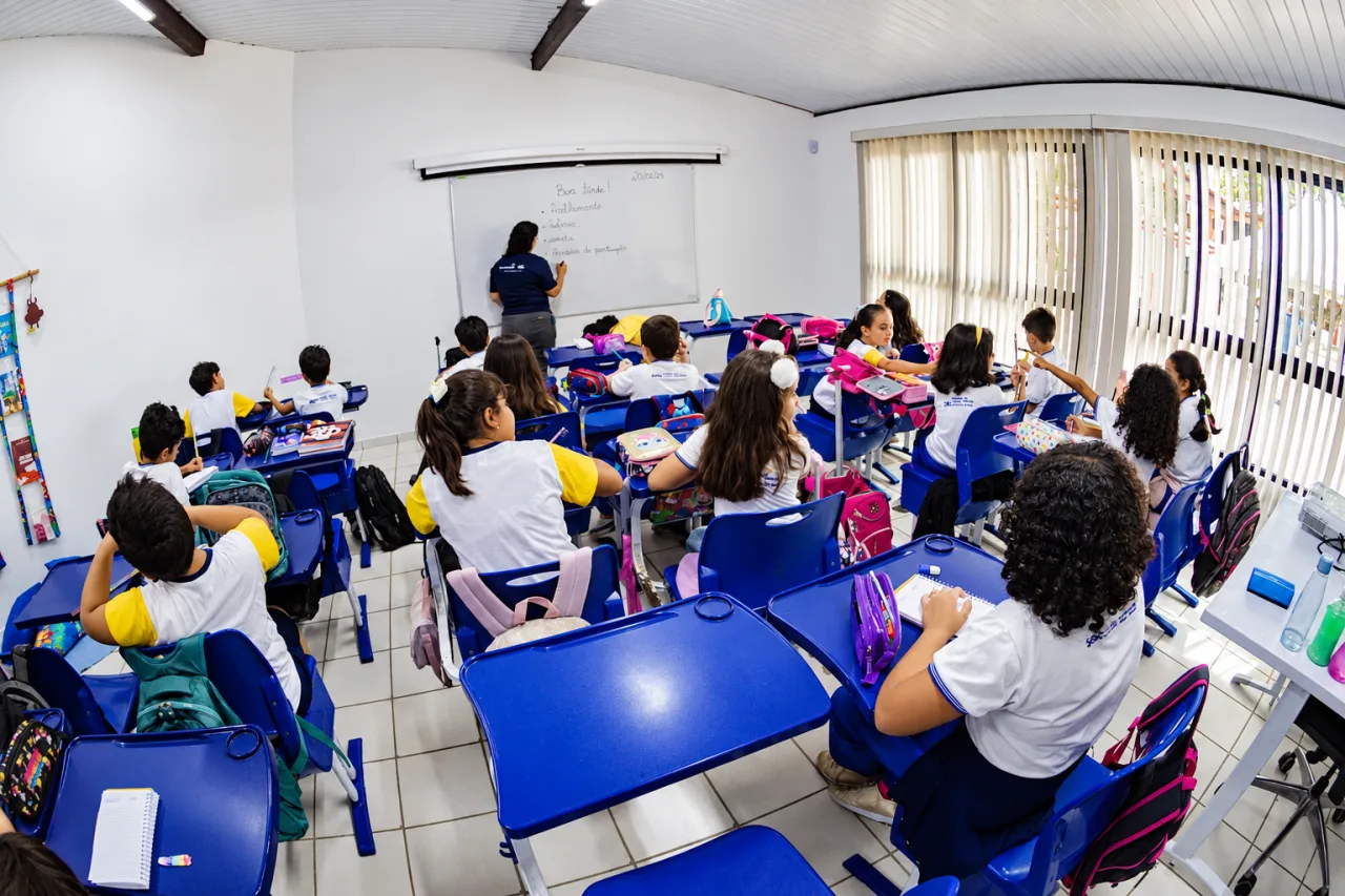 Estudantes uniformizados sentados em carteiras azuis acompanham professora diante do quadro