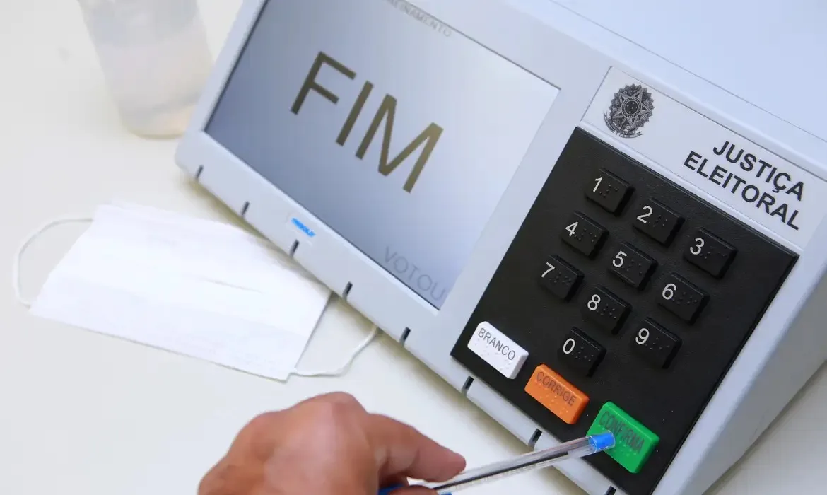 Urna eletrônica sendo substituída por equipe técnica em seção eleitoral.
