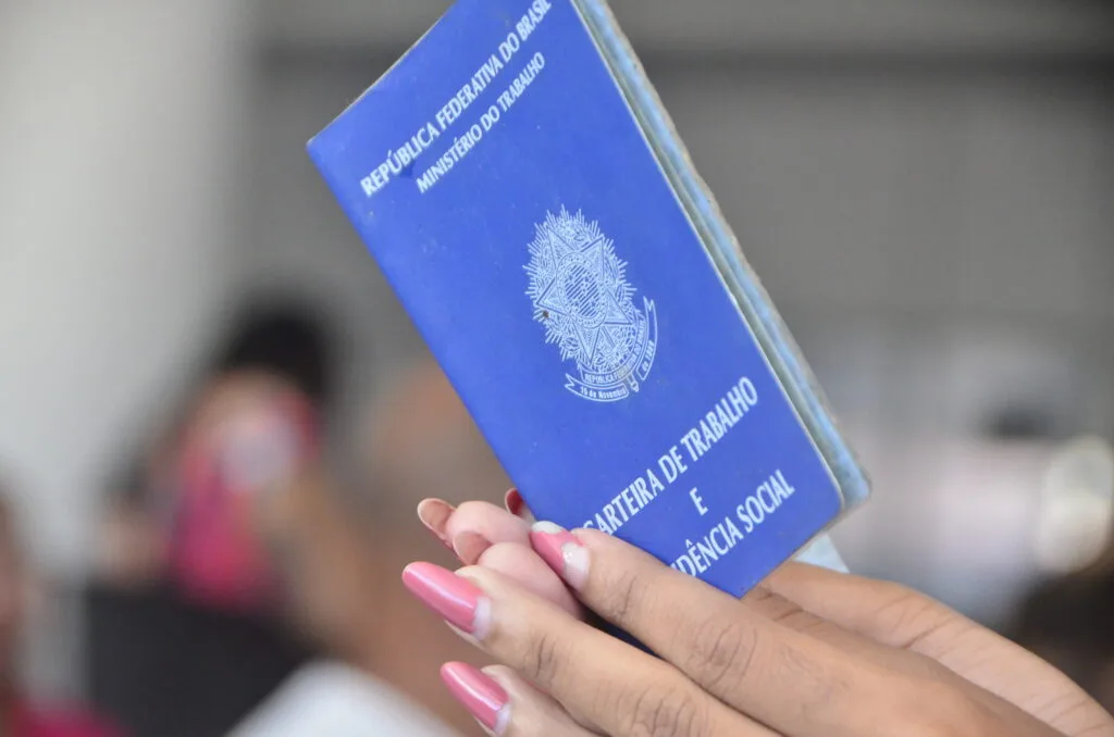 Pessoa segura uma carteira de trabalho azul diante da câmera